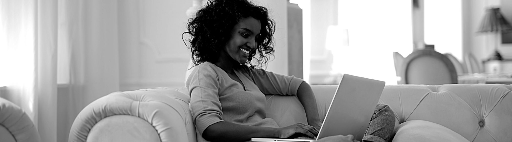 image depicts a woman at home on her sofa, working on a laptop and smiling